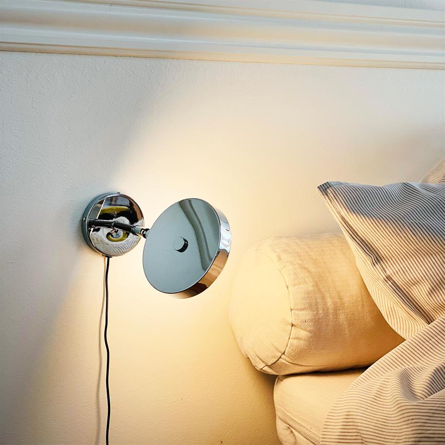 En krom væglampe kaster varmt, fokuseret lys; den er monteret over en beige sofa med rund nakkepude og stribet pude i et lyst, roligt soveværelsesmiljø.