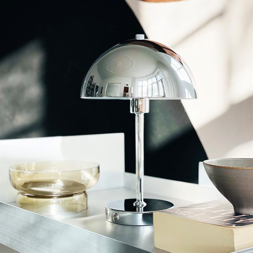 En krom bordlampe reflekterer rummet, står på en hvid bakke ved en transparent skål og en bog; blødt lys og kontrasterende mørk bagvæg i et lyst, moderne interiør.