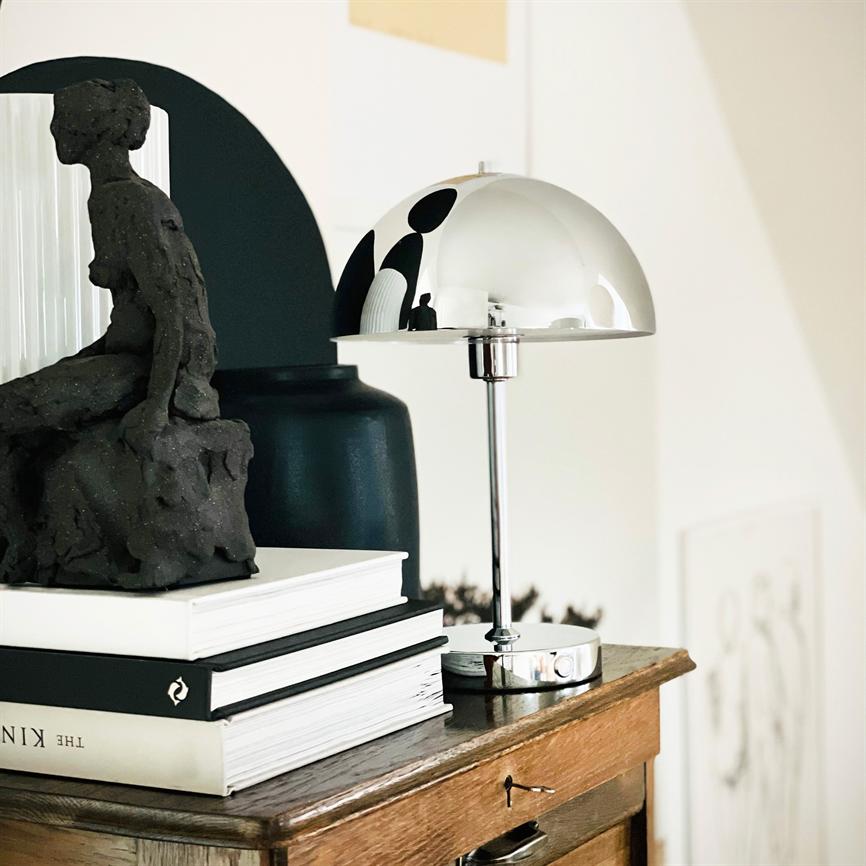 En krom bordlampe reflekterer omgivelserne; den står ved siden af en mørk skulptur på en stak bøger ovenpå et træskab i et lyst, minimalistisk rum.
 
 Tekst: "THE KIN"
