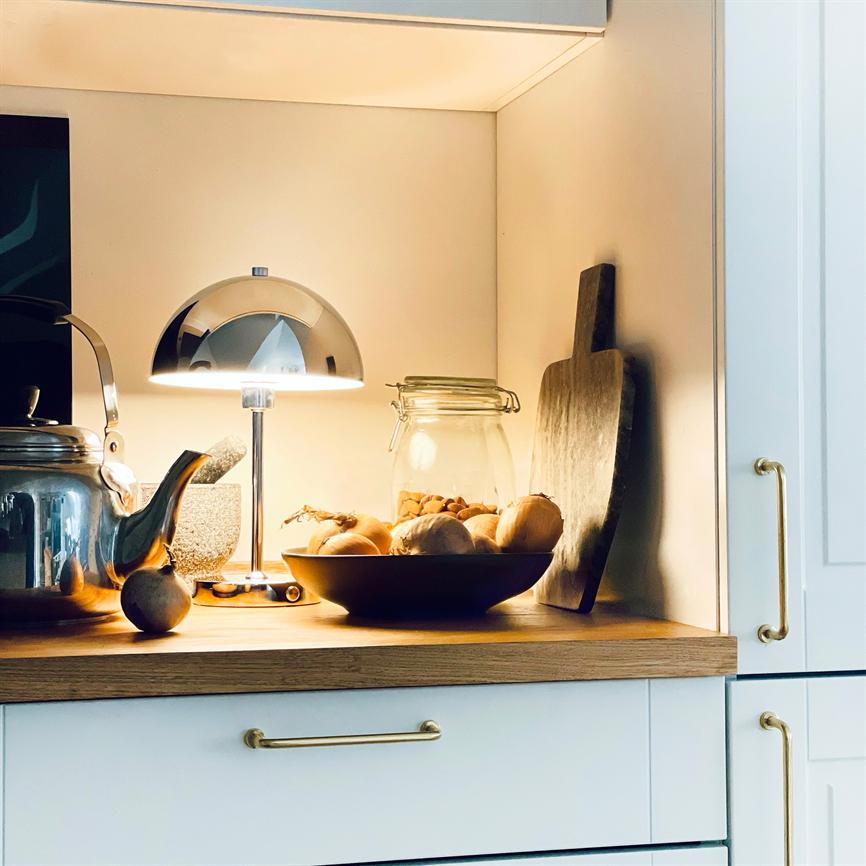 En poleret metalbordlampe lyser over en skål med løg; et glas og en tekande står ved siden af på en træbordplade i en oplyst, moderne køkkenniche.