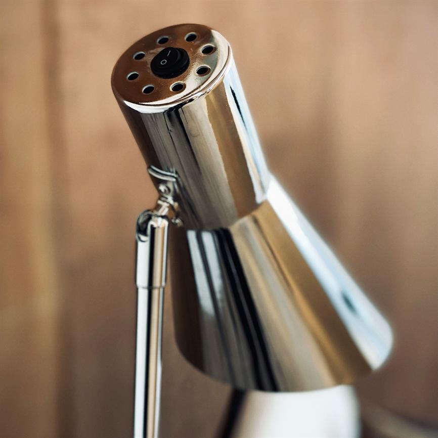 En krom bordlampe — lampehovedet er vinklet frem og reflekterer lys, med ventilationshuller og sort kontakt mærket "I" — foran en sløret, lys træbaggrund i nærbillede.