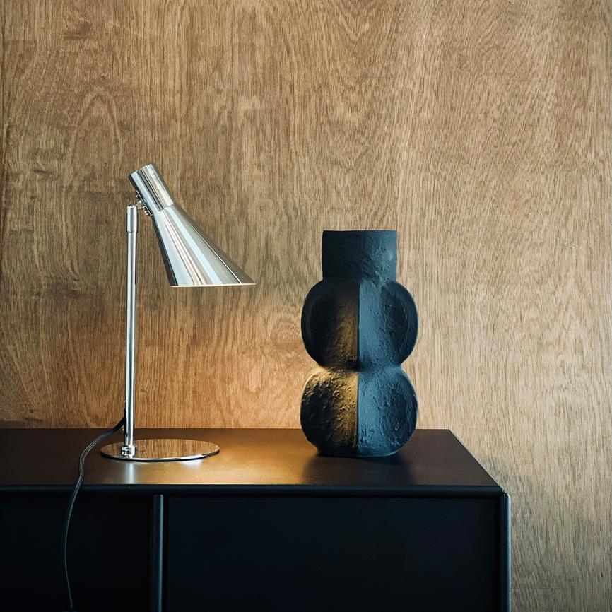 Skrivebordslampe lyser svagt mod en skulpturel, sort vase; begge står på et mørkt skab foran en varm, træbeklædt væg.