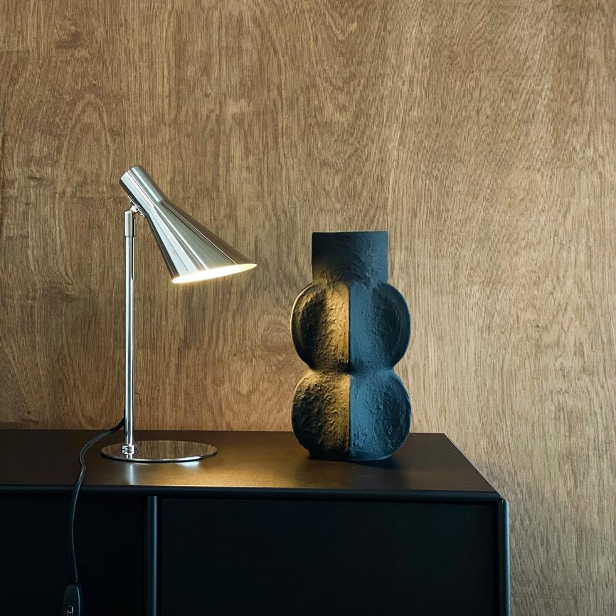 Metal bordlampe lyser på en sort, tredelt skulpturel vase placeret på et sort skab foran en lys træbeklædt væg, hvilket skaber kontrast mellem varme og kølig tekstur.