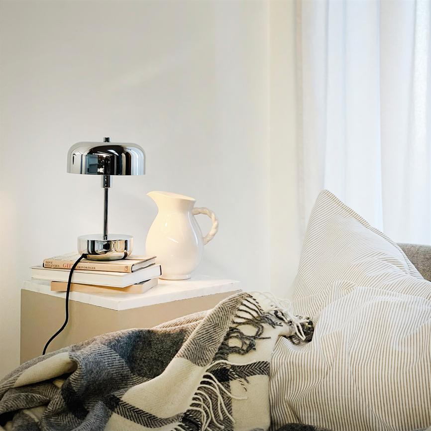 Krom bordlampe står på et natbord ved siden af en hvid kande; en stak bøger ligger under lampen; stribet pude og ternet plaid danner en hyggelig, lys soveværelseskrog.