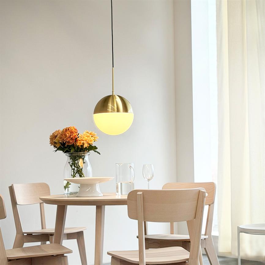En rund gylden pendellampe lyser over et rundt træbord med fire lyse træstole; bordet har en glasvase med orange blomster, en karaffel, et vinglas og lyse gardiner i baggrunden.