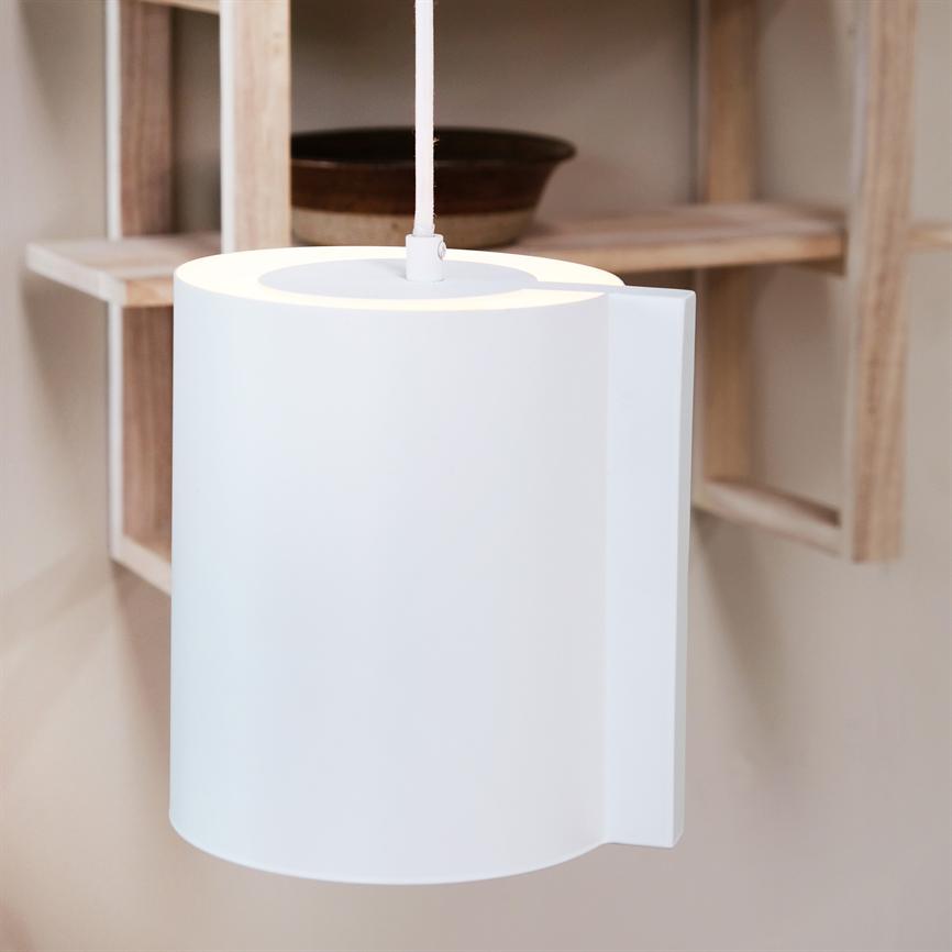 Hvid cylindrisk pendellampe hænger og udsender blødt lys fra toppen foran en åben træhylde med en keramisk skål på en beige væg.
