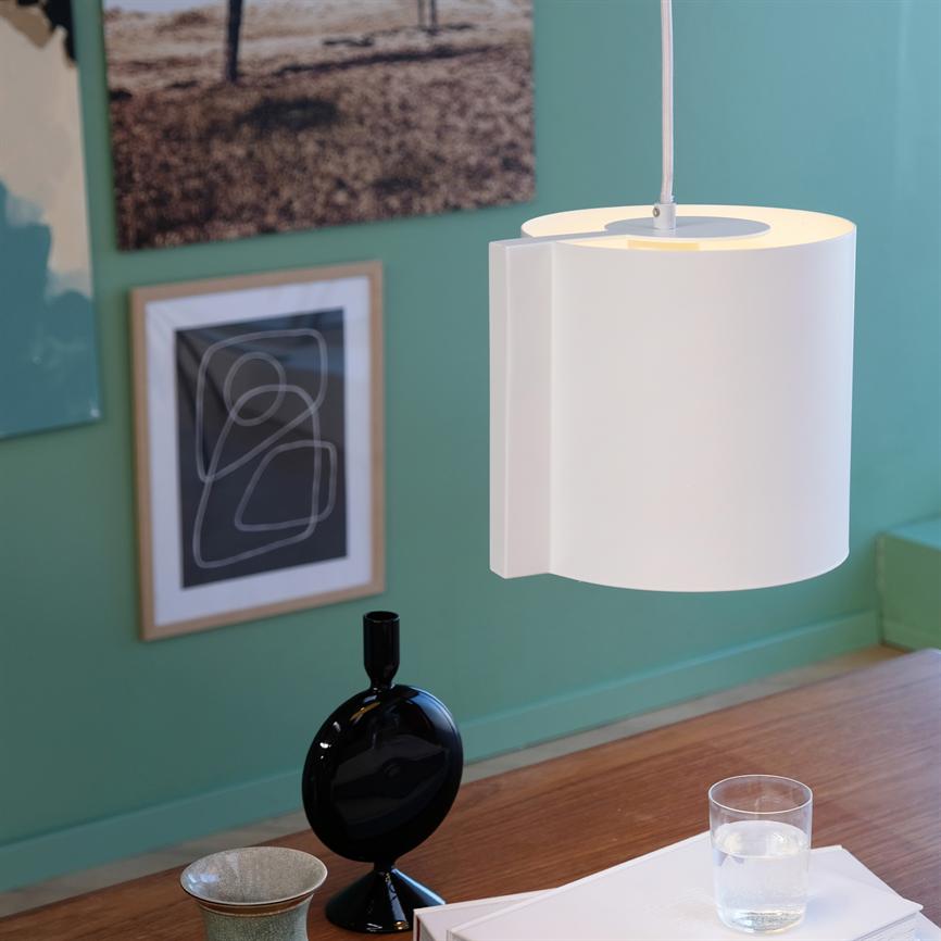 En hvid pendel-lampe hænger og kaster blødt lys over et træbord; på bordet står en sort vase, et glas vand og et par papirer foran en grøn væg med indrammede plakater.