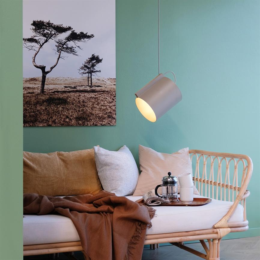 En rattan-sofa med puder og et brunt tæppe står i forgrunden; en hængelampe lyser over en bakke med fransk presse og kopper. Mintgrøn væg og et fotografi af træer i baggrunden.