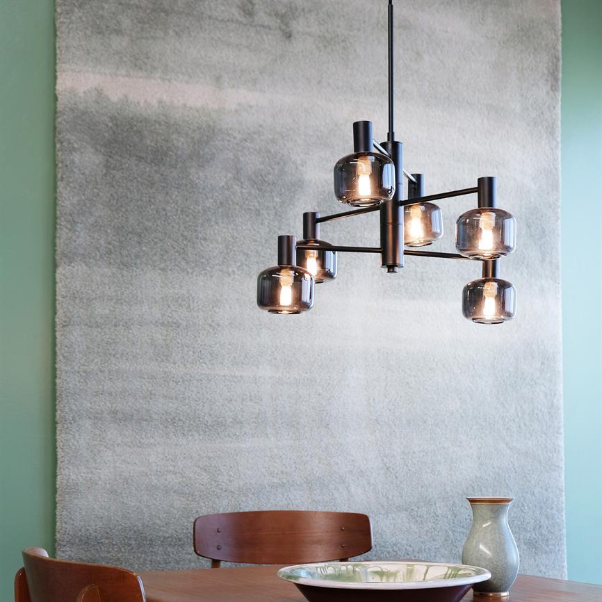 En modern loftlampe med seks røgfarvede glaskupler, der kaster varmt lys over et lyst træbord; omgivet af to træstole, en skål, en vase og et stort lysegråt væg‑tæppe.