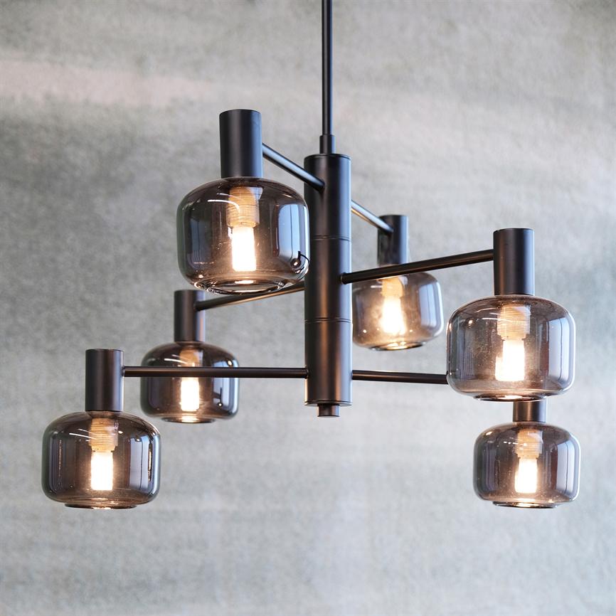 En moderne loftslampe med seks røgfarvede glaskupler, hvor pærerne lyser svagt, ophængt i sorte metalarme foran en tekstureret lysegrå væg.
