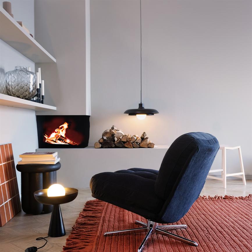Sort loungestol står lidt vendt mod en ild i pejsen i en moderne, minimalistisk stue med stak brænde, hængelampe, lille bord med rund lampe og rødt tæppe med frynser.