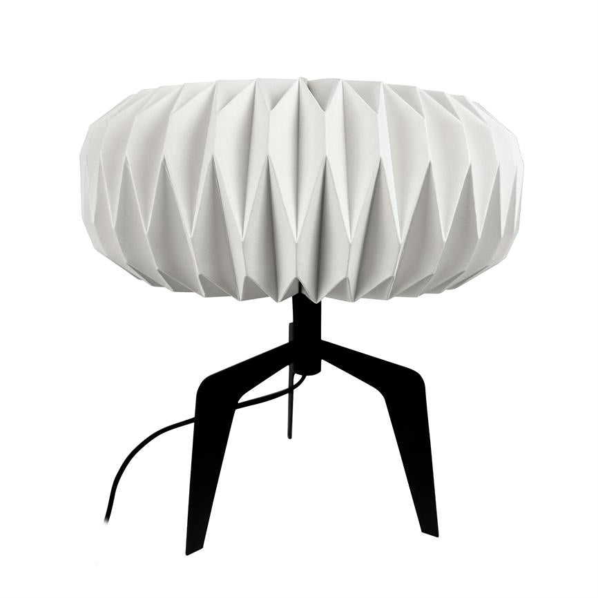 Bordlampe står på en sort trefod og har en rund, hvid origami‑foldet skærm; sort ledning hænger ned mod en neutral hvid baggrund.