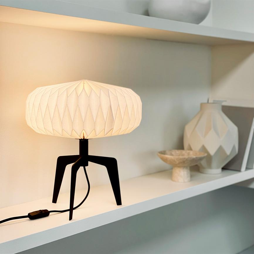 Bordlampe står tændt på en trebenet sort fod, kaster varmt lys gennem en foldet hvid skærm; dekorative keramikvaser og en lille skål står ved siden af på en hvid hylde.