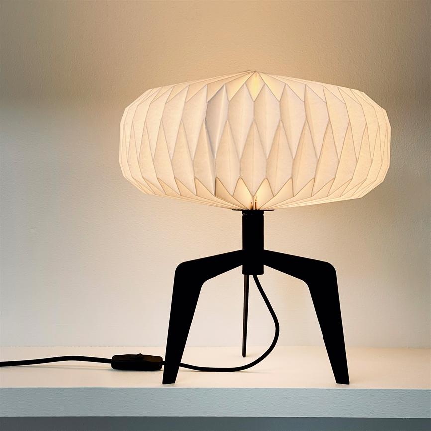 Bordlampe med origami-lignende hvid skærm lyser blødt; sort trebenet stativ står på en hvid hylde mod en lys væg, synlig ledning.