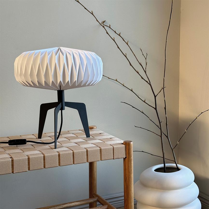 Lampe står på en flettet træbænk; hvid, geometrisk skærm på sort trefod. Tynde, nakne grene stikker op fra en hvid rund vase mod en neutral væg.