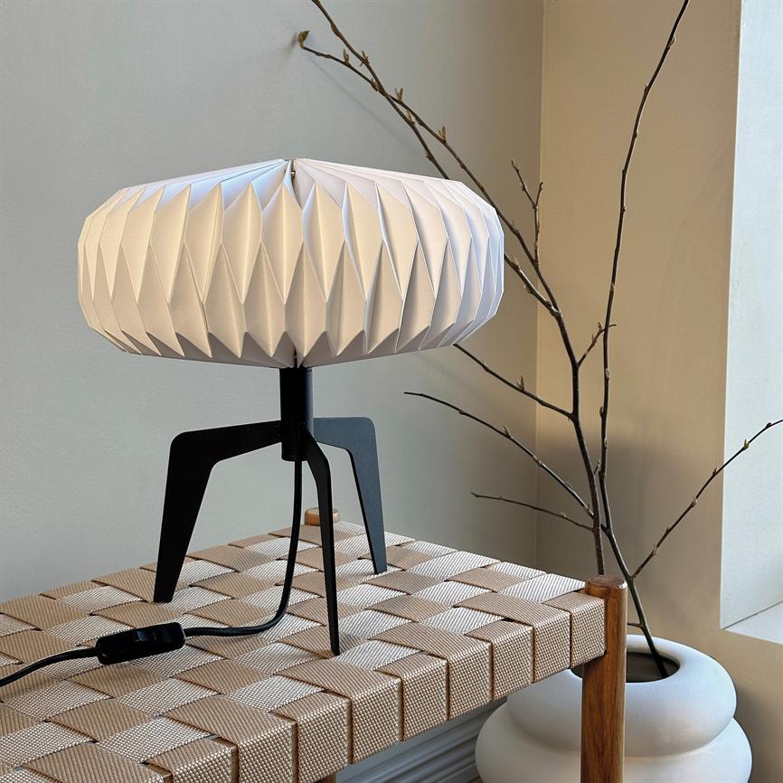 En hvid, foldet bordlampe på sort trefod står på en beige flettet bænk; en sort ledning ligger foran, og tørre grene i en hvid vase hviler ved vinduet.