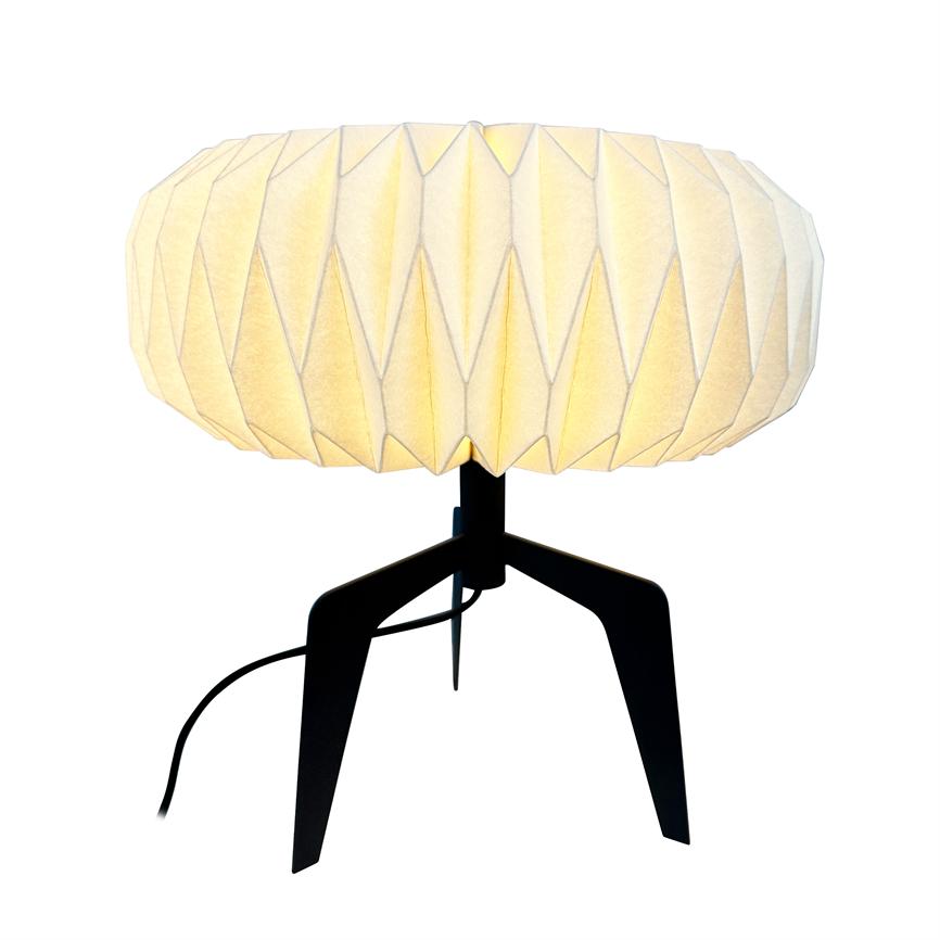 Bordlampe med rund, foldet papirskærm, der udsender et varmt, diffust lys; står på sort trefod med synlig ledning, isoleret mod en hvid baggrund.