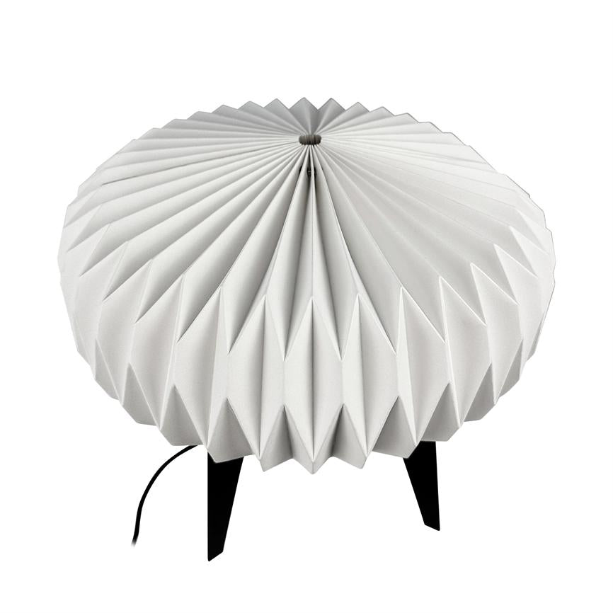 Rund lampe med plisseret hvid skærm hviler på tre sorte ben; på ren hvid baggrund ses et sort strømkabel løbe ud fra skærmens underkant.