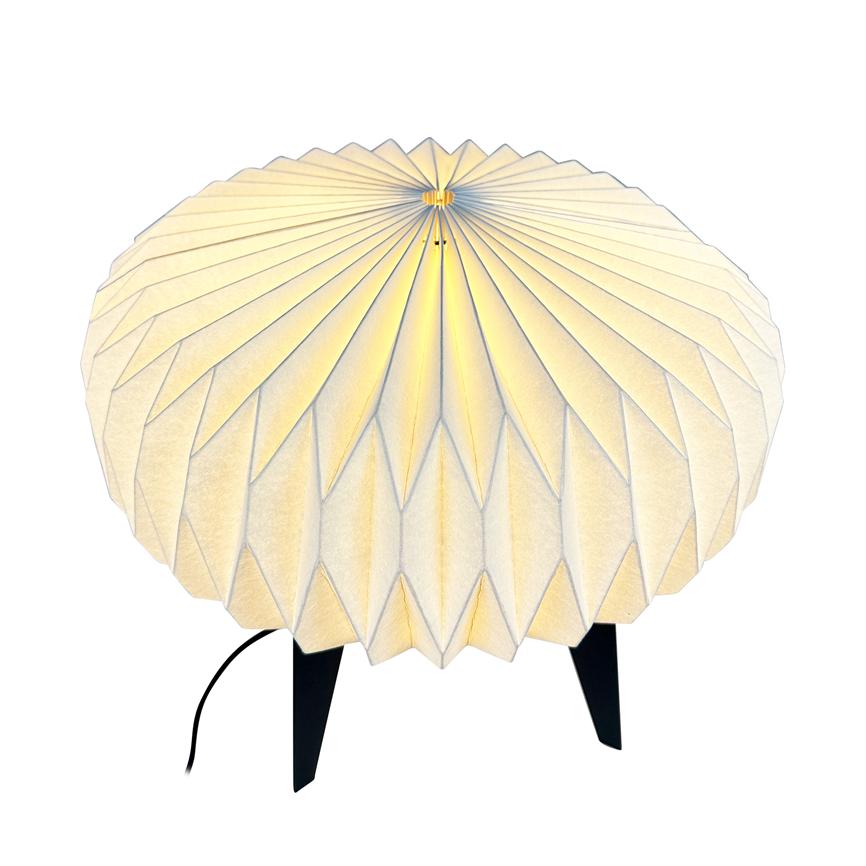 Rund, foldet papirlampe med rillede, radiære folder lyser blødt gult, står på tre sorte ben med synlig ledning på en neutral hvid baggrund.