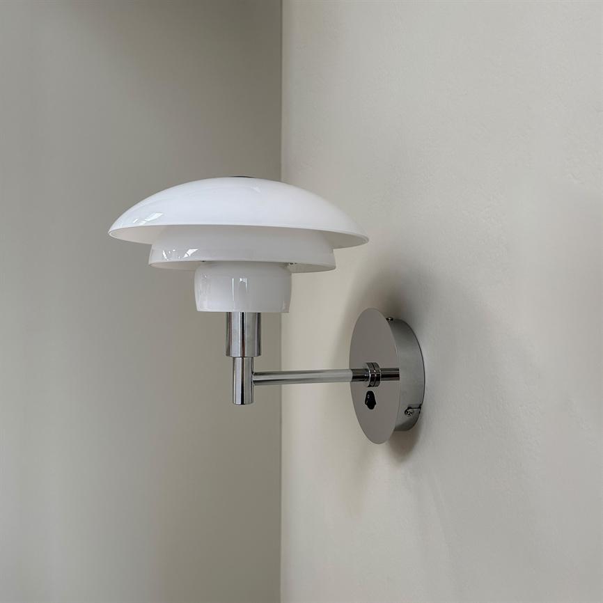 Væglampe monteret på en rund kromplade; en kort arm bærer en hvid, lagdelt glasskærm, som vender ud mod et lyst, blødt malet væghjørne.