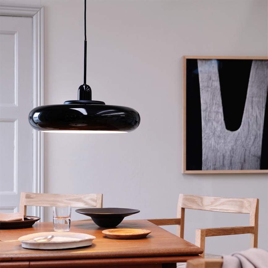 Sort pendellampe hænger over et træspisebord; keramiske fade, et vandglas og lyse træstole står klar i en lys, minimalistisk spisestue med et abstrakt sort-hvidt billede på væggen.