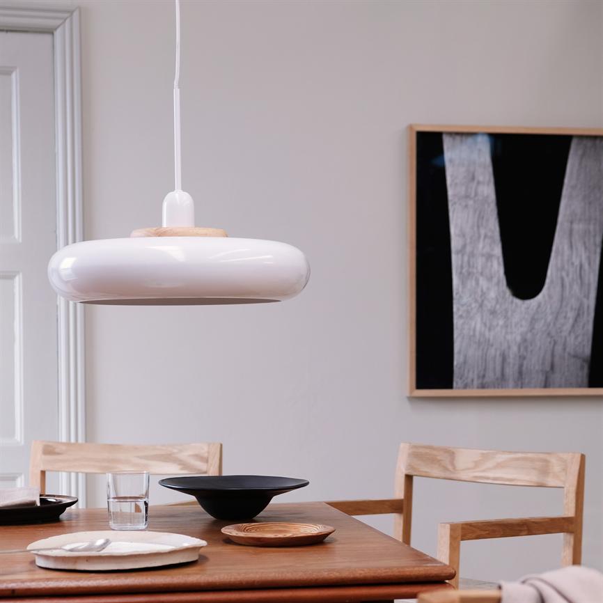 Rund hvid pendellampe hænger over et træspisebord med skåle, tallerkener og et glas; to træstole og et abstrakt sort-hvidt billede pryder væggen i en lys, minimalistisk spisestue.