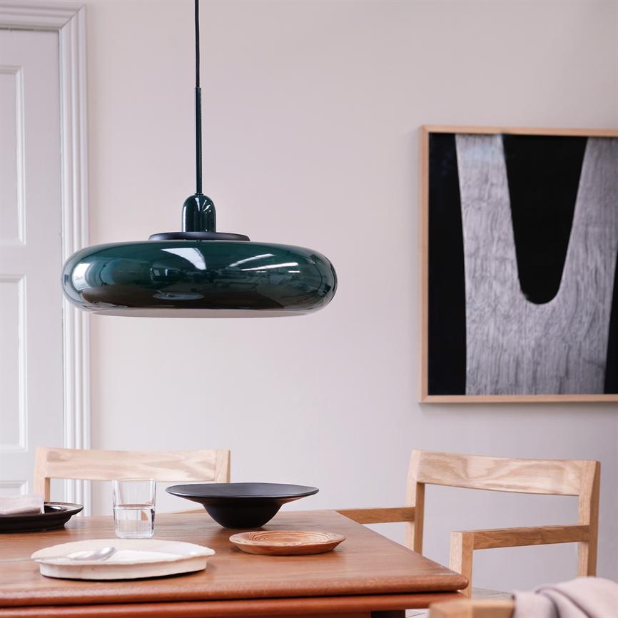 En blank grøn pendellampe hænger lavt over et træspisebord; bordet er dækket med tallerkener, skåle og et glas, omgivet af lyse træstole i et minimalistisk rum med abstrakt vægkunst.