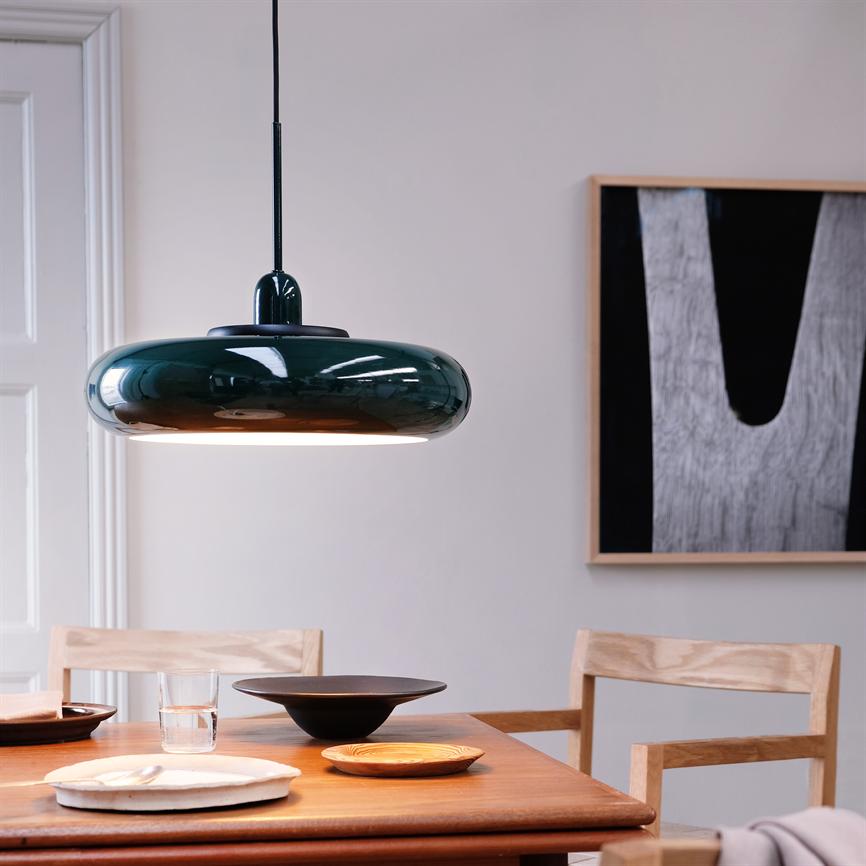 En mørkegrøn glaseret pendel lyser ned over et træbord med tallerkener, skåle og et glas; omgivet af lyse træstole og abstrakt sort-hvidt vægkunst i et lyst, minimalistisk rum.