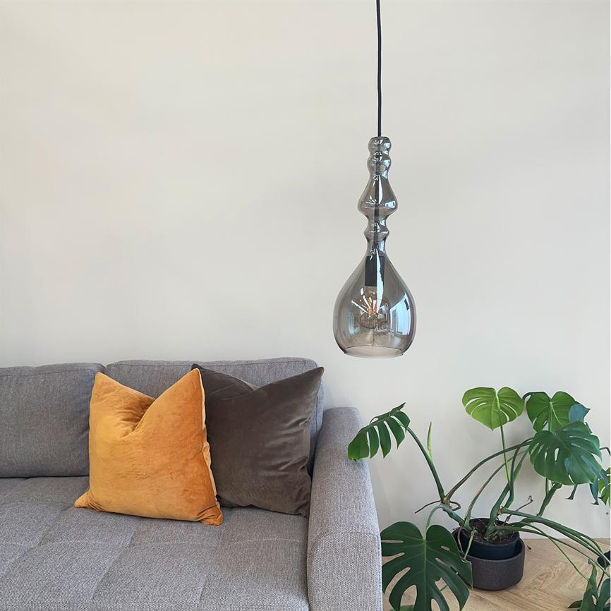 En dekorativ hængende glaspære svæver over en grå sofa; to velourpuder (gul og brun) ligger på sofaen, og en stor monstera-plante står i potte på trægulvet.