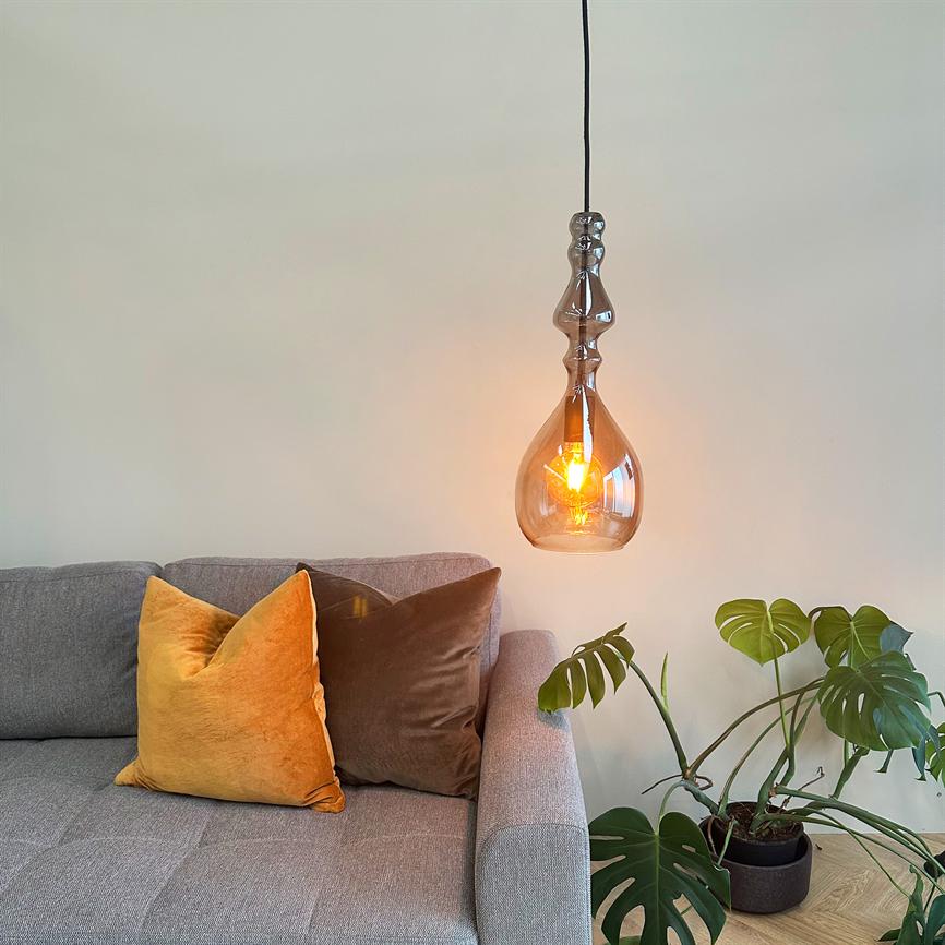 Hængende glaspære lyser varmt over en grå sofa; to dekorative puder (orange og brun) ligger på sofaen, og en grøn monstera-plante står i en potte på trægulvet.