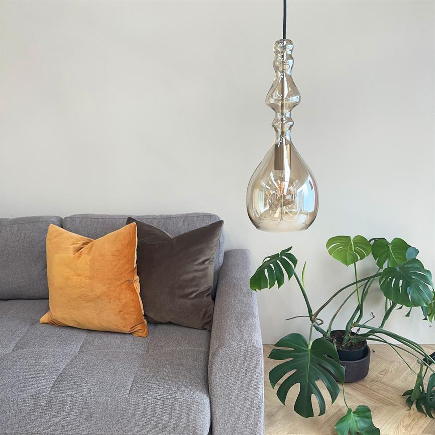 En dråbeformet glaspendel hænger over en lysegrå sofa og kaster blødt lys; sofaen har en gul og en brun pude, og en stor Monstera i potte står på lyst trægulv.