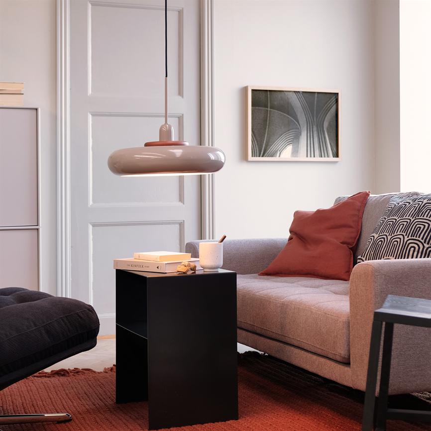 Lille sort sidebord står med en kop og en bog mærket "KINFOLK" foran en grå sofa i en lys stue med hængende rund lampe, rødbrunt tæppe og vægkunst.