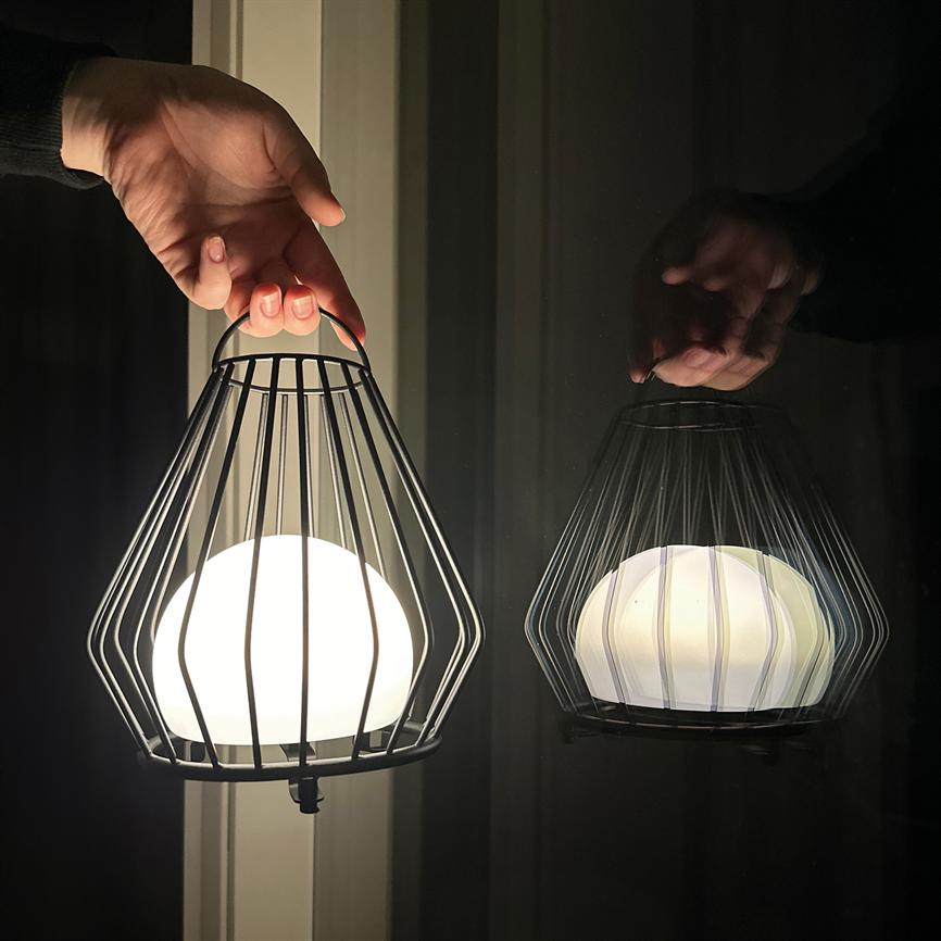 En kugleformet lampe indkapslet i et metalgitter holdes i et håndtag; den udsender varmt lys, og både lampe og hånd spejles i en mørk vinduesrude.