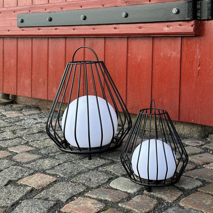 To sorte metallanterner, én stor og én mindre, holder hvide kuglelamper foran en rød træbygning, stående på et brostensbelagt underlag.