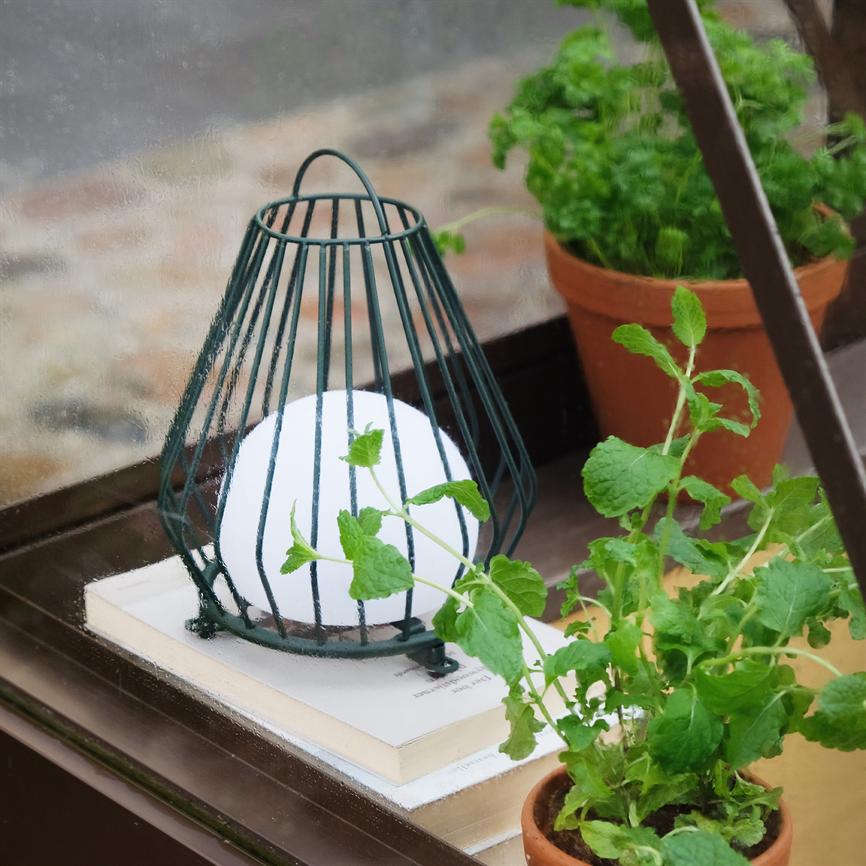 En rund hvid lampe i et grønt metalbur står på en stak bøger på en vindueskarm, omgivet af terrakottakrukker med grønne krydderurter og en regnvåd rude.