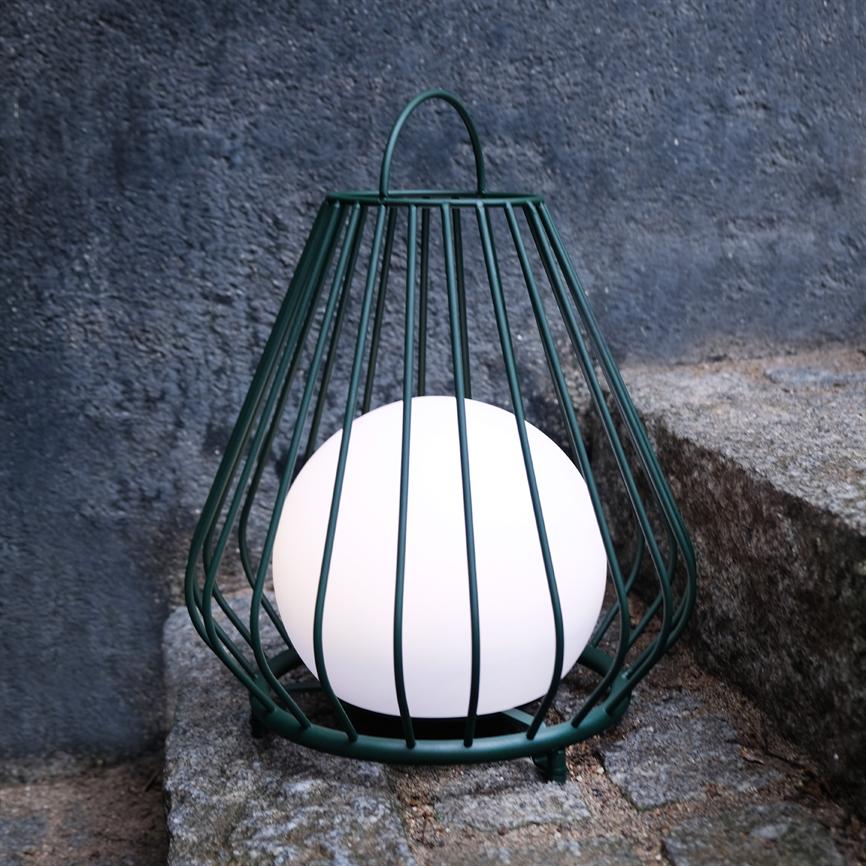 En kugleformet lampe lyser inde i et mørkegrønt metalbur, placeret på ru granittrin udendørs foran en grå, tekstureret betonvæg.