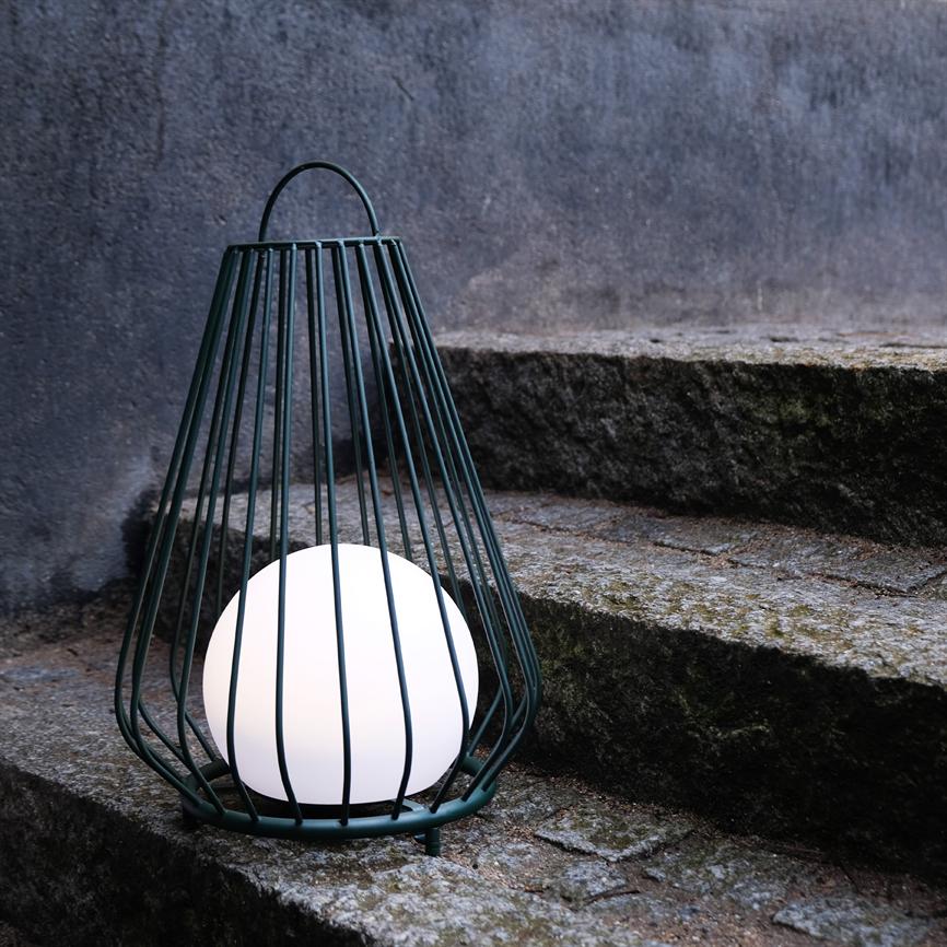 En hvid kugleformet lampe i et mørkegrønt metalgitter lyser blødt og står på mosdækkede, vejrbidte stenetrin foran en grå, ru væg.