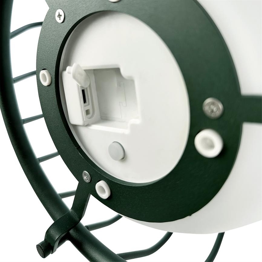 Rund hvid plastdisk viser en åben batteri- og USB-port; en mørkegrøn metalramme omkranser den med synlige skruer og gummipuder — nærbillede af bagenden af en lille enhed.