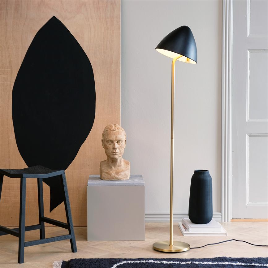 Gulvlampe kaster varmt, nedadrettet lys over et minimalistisk hjørne med en stenskulptur på en piedestal, en høj sort vase, en sort træskammel og en stor træplade med sort oval.
