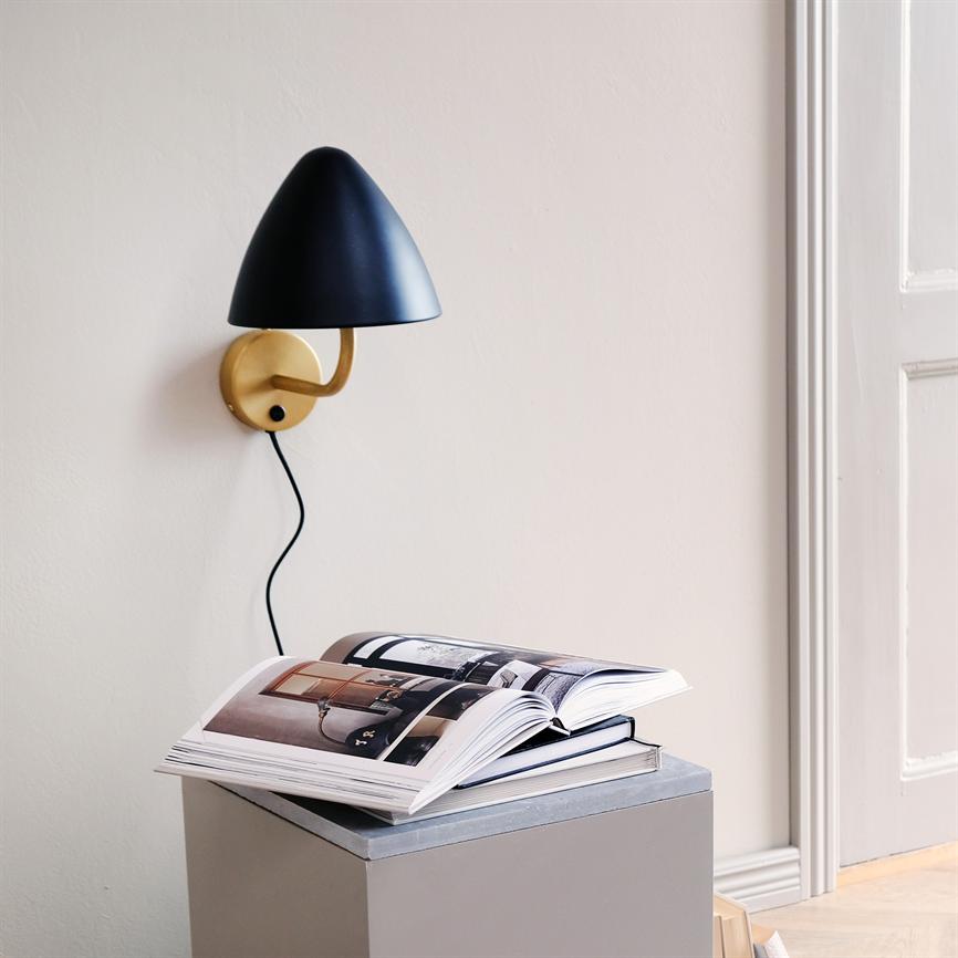 Væglampe med sort skærm og guldfarvet arm hænger med synlig ledning langs en lys væg. Et åbent fotobog ligger på en grå kasse under lampen, ved en dørkarm.