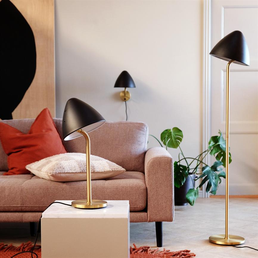 Sort og guldfarvet bordlampe står på et hvidt sidebord; en pudderrosa sofa med røde og lyse puder bagved; matchende gulv- og væglampe samt grøn stueplante i et lyst hjørne.