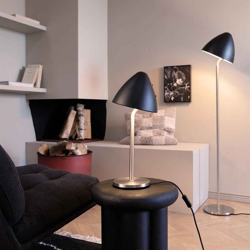 Sort bordlampe med buet skærm står og lyser blødt på et rundt sort sidebord, foran en mørk sofa; bagved står matchende gulvlampe, hvid skænk med tæppe og brændekurv.