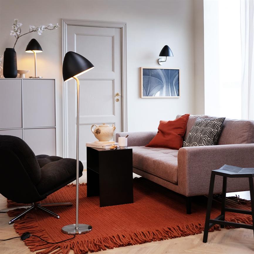 Sofaen står ved et vindue, flankeret af et lille sort bord og en tændt gulvlampe; farvede puder ligger på den, over et rødt frynset tæppe i et lyst, moderne stuemiljø.