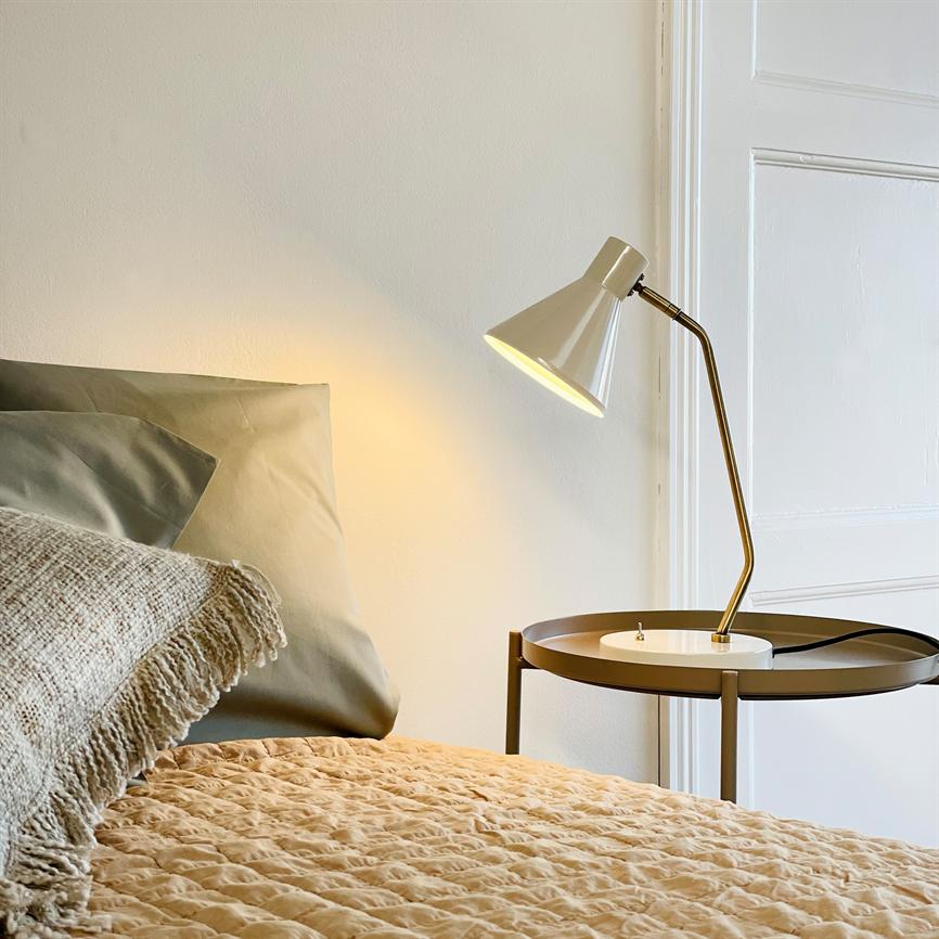 Bordlampe kaster varmt lys over seng; står på rundt metallisk natbord ved en hvid dør; foran quiltet beige sengetæppe og en tekstureret grågrøn pude.