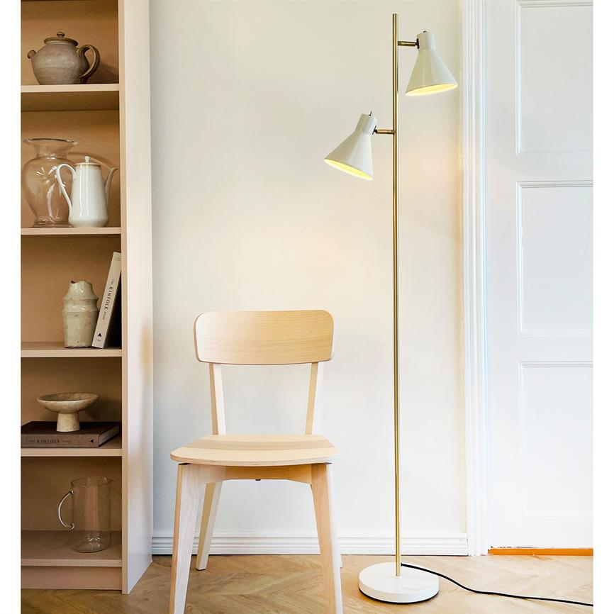 Træstol står stille foran en hvid væg; en toarmet gulvlampe lyser ved siden af, og en åben reol med keramik og bøger står til venstre i et lyst, minimalistisk rum.