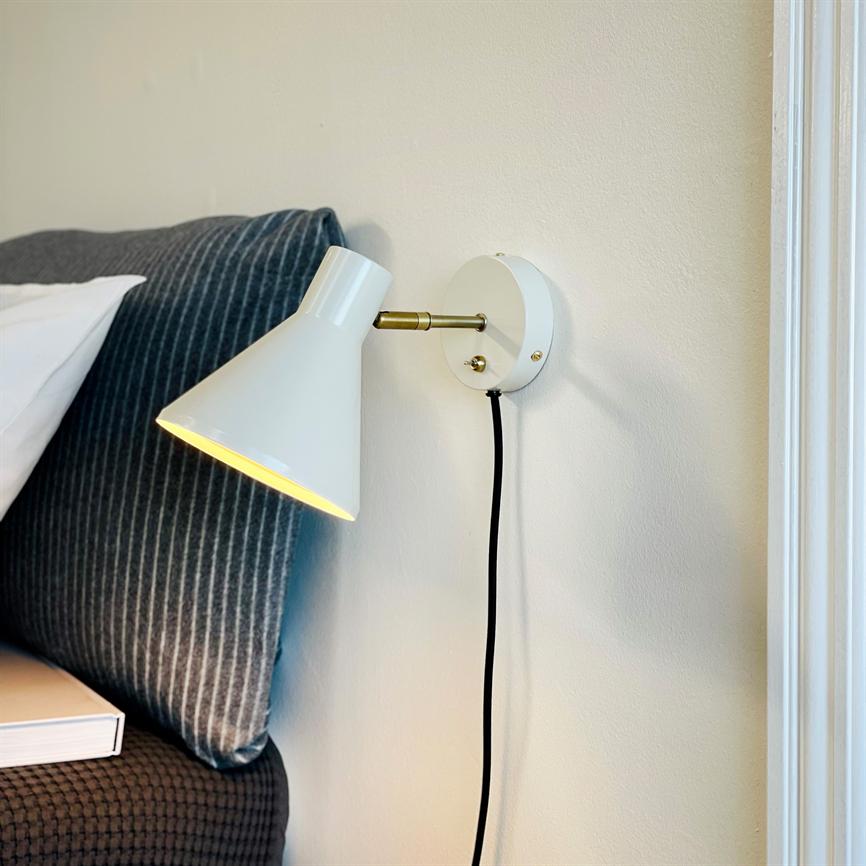 Hvid væglampe lyser og kaster varmt lys ned mod sengegavl; monteret på lys væg med sort ledning, ved en seng med mørkegrå, stribet pude og en bog på sengekanten.