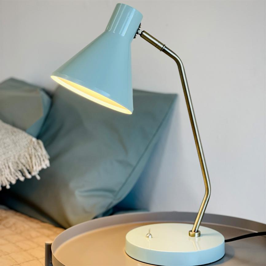 En blå metal bordlampe lyser nedad fra en buet messingarm; den står på et rundt natbord ved en seng med gråblå puder og et flosset, hvidt kastetæppe.