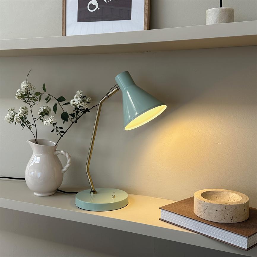 En mintgrøn skrivebordslampe lyser varmt nedover en væghylde, flankeret af en hvid kande med hvide blomster til venstre og en bog med en rund stenskål til højre.