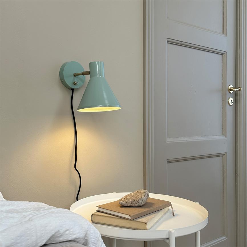 En mintgrøn væglampe lyser blødt ned over et rundt hvidt sidebord med to bøger og en sten, ved siden af en seng og en lysegrå paneldør på en beige væg.