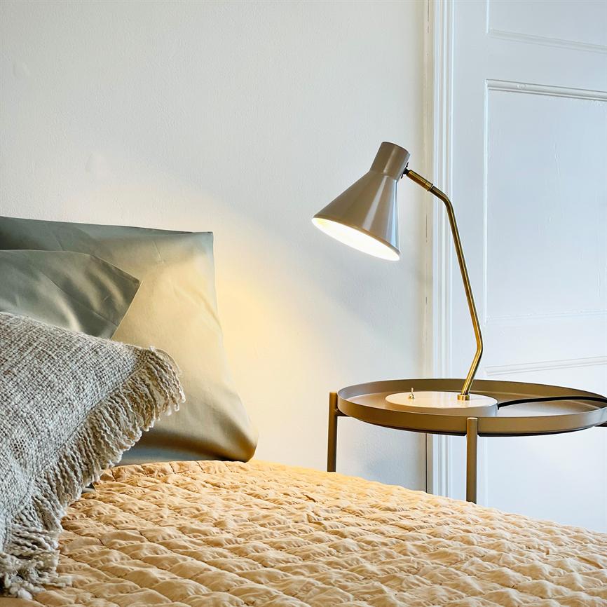 En bordlampe lyser varmt ned over et rundt natbord; den kaster lys på en quiltet beige dyne og en grøn pude, i et enkelt, lyst soveværelse med hvid dør.